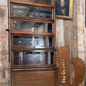 May include: An antique wooden barrister bookcase with glass-fronted shelves. The bookcase has multiple sections with pull-out shelves and a dark wood finish. A framed portrait hangs on the wooden wall behind the bookcase.