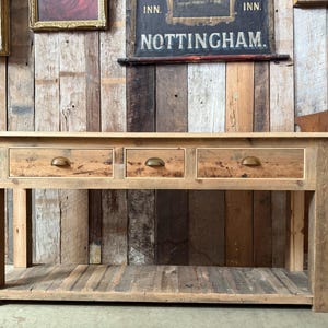 May include: A rustic wooden console table with three drawers and a lower shelf. The table is made of reclaimed wood, with visible knots and grain. The drawers have brass-coloured pulls. The table is approximately 152 cm long, 46 cm deep, and 76 cm high.