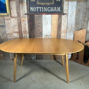 May include: Oval wooden dining table with four tapered legs. The table has a light brown finish and a smooth surface. The table is approximately 152 cm long and 102 cm wide.