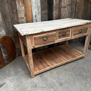 May include: A rustic wooden console table with three drawers and a lower shelf. The table has a light-coloured top and a natural wood finish. The drawers have metal pulls. The table is approximately 122 cm long, 46 cm wide, and 76 cm high.