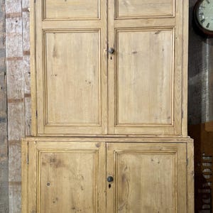 May include: A tall, two-part wooden cabinet in a light brown hue. The upper section features two doors, and the lower section also has two doors. Each door is fitted with a dark knob. The cabinet is likely for storage.