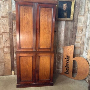 May include: A tall, antique wooden cabinet with four doors, in a warm brown colour. The cabinet is set against a rustic wooden wall, with a framed portrait and a sign that reads "YORKEY" above it. A large, decorative "b" is also visible.