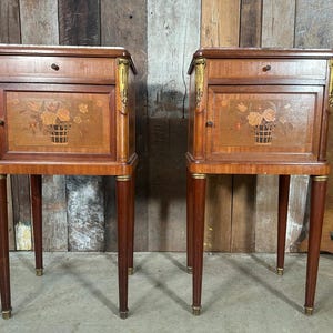 May include: Two antique wooden bedside cabinets. Each cabinet has a marble top, a drawer, and a door with a floral basket inlay. The cabinets have slender, tapered legs with brass accents and decorative gold trim.
