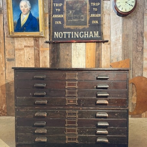 May include: A dark wooden flat file cabinet with multiple drawers. The cabinet has metal hardware and is set against a rustic wooden wall. Above the cabinet is a vintage sign that reads "Yorkey Trip to Jerusalem Inn Nottingham."
