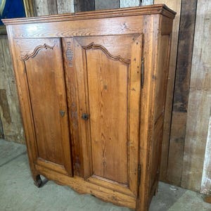 May include: An antique wooden cabinet in light brown. The cabinet has two doors with a decorative, curved top edge and small, dark knobs. The wood grain is visible. The cabinet has a distressed appearance, suggesting age and use. The cabinet's height is approximately 150 cm.