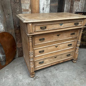 May include: A light-coloured wooden chest of drawers with four drawers and ornate detailing. The chest has a rectangular top and brass hardware. The wood has a natural finish, showing the grain.