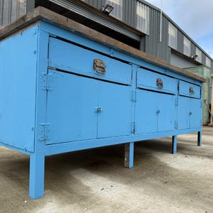May include: A large, vintage blue workbench with a weathered wooden top. The workbench has multiple drawers and cabinet doors with metal hardware. The legs are sturdy and painted the same blue as the body. A rustic, functional piece.