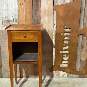 May include: A small, antique wooden nightstand with a single drawer and an open shelf below. The nightstand is made of light brown wood and has a simple, elegant design. The legs are slender and tapered. A rusty metal sign with the word "belvoir" is in the background.