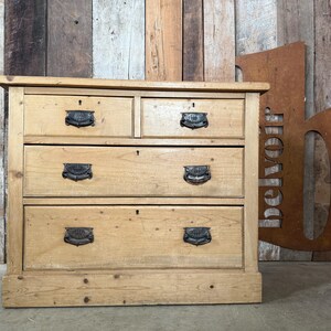 May include: A wooden chest of drawers in a light wood finish, featuring four drawers with ornate metal handles. The chest is set against a backdrop of wooden planks, creating a rustic aesthetic.