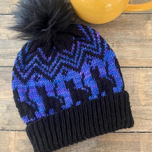 May include: A black knitted beanie hat with a blue and purple geometric pattern and a black faux fur pom-pom. A yellow teapot is in the background. The hat has a ribbed brim.