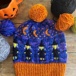 May include: A hand-knitted beanie featuring a vibrant orange ribbed brim and a matching orange pom-pom on top. The main body displays a purple and blue variegated background with a repeating pattern of black witch silhouettes and orange crescent moons.