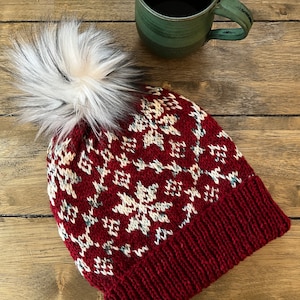Puede incluir: Un gorro de punto rojo con un pompón blanco y un patrón de copo de nieve. El gorro está sobre una superficie de madera.