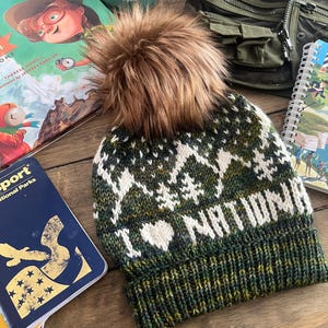 May include: A green, white, and brown knitted beanie with a faux fur pom pom. The beanie has a white knit pattern that says "I LOVE NATIONAL PARKS".