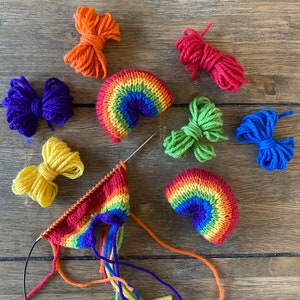 Könnte beinhalten: Ein Regenbogen-Strickprojekt mit Garn in verschiedenen Farben, darunter Rot, Orange, Gelb, Grün, Blau, Indigo und Violett. Das Garn ist in einem Kreis um eine Holzoberfläche angeordnet, wobei einige Garnknäuel noch in ihrer Originalverpackung sind.