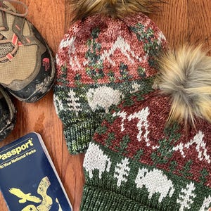 May include: Two knitted beanies with faux fur pom-poms, featuring a mountain and animal pattern in red, green, and white. A hiking shoe and a blue "Passport to Your National Parks" book are also visible.