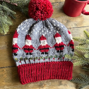 May include: A knitted winter hat in grey and red, topped with a red pom-pom. The hat showcases a pattern of Santa Claus figures and a red ribbed brim. It's displayed on a wooden surface, alongside a red mug and evergreen sprigs.