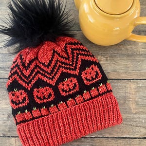 Op de afbeelding: Een rood-zwarte gebreide muts met een zwarte imitatiebont pompon. De hoed heeft een herhalend patroon van pompoenen en zigzags. Een gele theepot staat op de achtergrond.