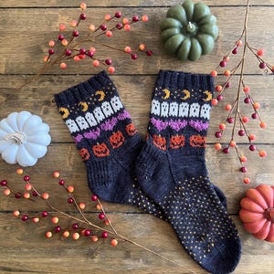 May include: A pair of black knitted socks with a Halloween-themed design. The socks feature a pattern of ghosts, pumpkins, and hearts in shades of white, orange, pink, and yellow.