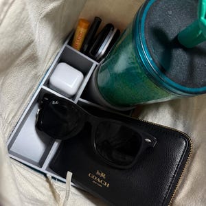 May include: A beige canvas tote bag containing a grey plastic organizer insert. The organizer holds a teal tumbler with a straw, a black Coach wallet, black Ray-Ban sunglasses, a white earbud case, and several small cylindrical items.