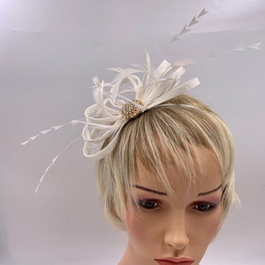 Pode incluir: Um pente de cabelo branco de penas e tecido com um pequeno enfeite redondo, brilhante e dourado.