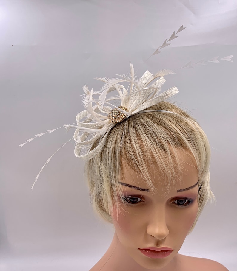 Pode incluir: Um pente de cabelo branco de penas e tecido com um pequeno enfeite redondo, brilhante e dourado.