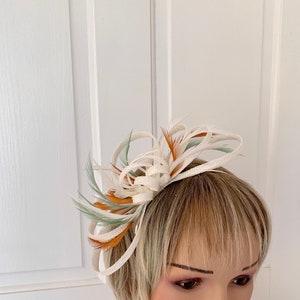 May include: A white feather fascinator with orange and green feathers. The fascinator is worn on a mannequin's head.