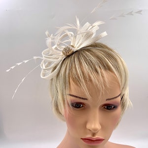 Pode incluir: Um pente de cabelo branco de penas e tecido com um pequeno adorno redondo dourado com pedras de strass transparentes.