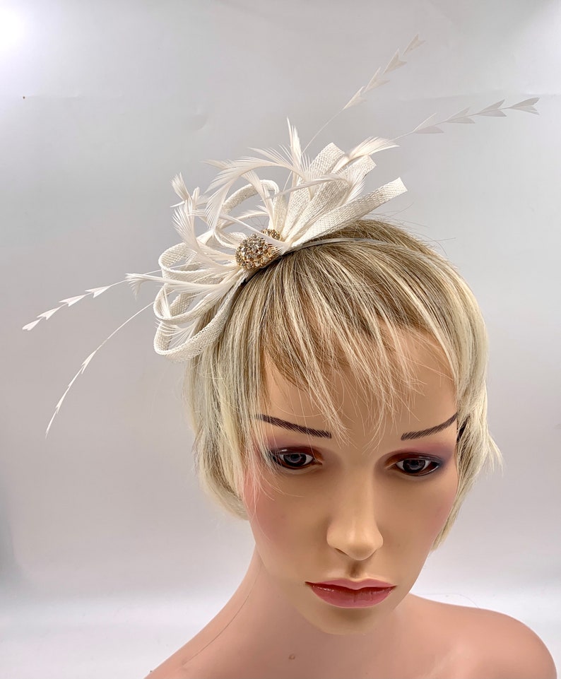 Pode incluir: Um pente de cabelo branco de penas e tecido com um pequeno adorno redondo dourado com pedras de strass transparentes.