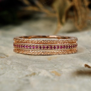 May include: A rose gold band ring featuring a central row of small, round, vibrant pink rubies. The ring is flanked by two decorative borders with an engraved geometric pattern and a beaded texture, creating a detailed, multi-layered appearance.