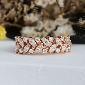 Puede incluir: Anillo de oro rosa con dise&ntilde;o de hojas y piedras blancas de talla marquesa y redonda. El anillo est&aacute; sobre una superficie blanca con un fondo floral borroso. Una fina joya.