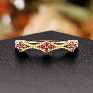 Op de afbeelding: Een goudkleurige ring met een delicaat ontwerp. De ring heeft kleine, ronde, rode edelstenen in een herhalend patroon. De ring ligt op een zwart oppervlak, met een vage bloemenachtergrond.