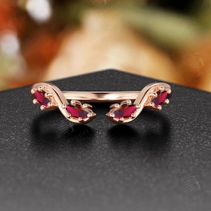 May include: Rose gold ring with a curved design. The ring features six oval-cut, deep red gemstones. The ring is set against a black surface with a blurred background of pink and yellow.