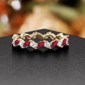 May include: A gold ring with alternating red and white gemstones. The ring features a repeating pattern of marquise-cut red rubies and round, clear diamonds. The ring is set on a black surface with a blurred floral background.