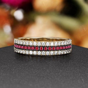 Pode incluir: Um anel de banda dourada com três fileiras de pedras preciosas. As fileiras superior e inferior são brancas, e a fileira do meio é vermelha. O anel está sobre um fundo desfocado rosa e verde.