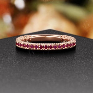 May include: Rose gold band ring with a row of small, vibrant red gemstones. The ring is set on a black surface, with a blurred floral background. The ring's design is simple yet elegant, showcasing the gemstones.