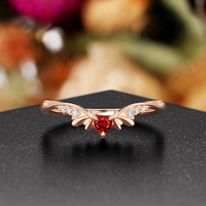 May include: Rose gold ring with a heart-shaped red gemstone center. The ring features a delicate design with small, clear accent stones. The ring is set against a dark surface with a blurred floral background.