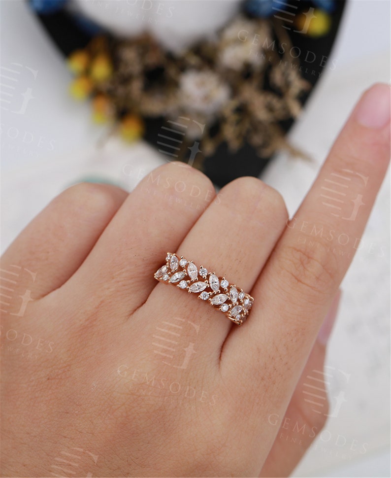 Puede incluir: Un anillo de oro rosa con una fila de piedras preciosas blancas con corte marquesa. El anillo est&aacute; engastado con m&uacute;ltiples piedras preciosas, creando un dise&ntilde;o brillante y elegante. El anillo se muestra en el dedo de una persona, destacando sus delicados detalles.