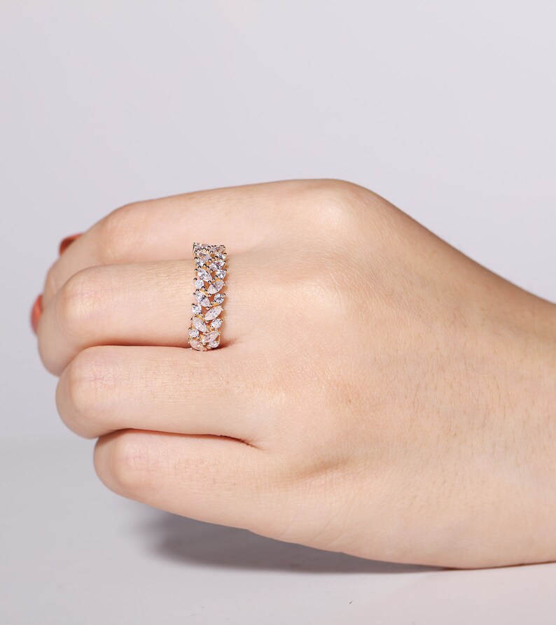 Puede incluir: Un anillo dorado con dos filas de piedras claras en forma de l&aacute;grima. El anillo se muestra en una mano, resaltando el dise&ntilde;o detallado y el brillo de las piedras preciosas. La banda del anillo tambi&eacute;n es dorada.