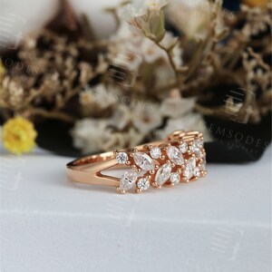 Puede incluir: Anillo de oro rosa con una doble fila de piedras preciosas blancas con corte marquesa y redondo. El anillo tiene un dise&ntilde;o de ca&ntilde;a dividida y est&aacute; engastado sobre un fondo borroso de flores blancas y vegetaci&oacute;n. Una joya fina.