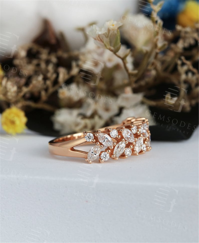Puede incluir: Anillo de oro rosa con una doble fila de piedras preciosas blancas con corte marquesa y redondo. El anillo tiene un dise&ntilde;o de ca&ntilde;a dividida y est&aacute; engastado sobre un fondo borroso de flores blancas y vegetaci&oacute;n. Una joya fina.