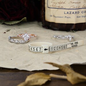 May include: Two rings are displayed with a plastic ring sizer. One ring features a kite-shaped center stone with side accents in rose gold, paired with a curved band. The other is a silver-toned ring with a marquise-cut stone and intricate detailing.