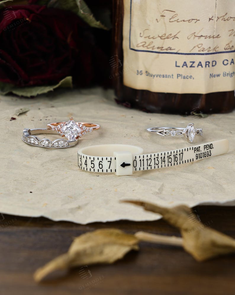 May include: Two rings are displayed with a plastic ring sizer. One ring features a kite-shaped center stone with side accents in rose gold, paired with a curved band. The other is a silver-toned ring with a marquise-cut stone and intricate detailing.