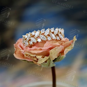 Puede incluir: Un anillo de oro con piedras preciosas claras de talla marquesa y redonda. El anillo est&aacute; sobre una rosa seca de color naranja claro. El fondo es una mezcla borrosa de azul, blanco y marr&oacute;n. El anillo es de Gemsodes Fine Jewelry.