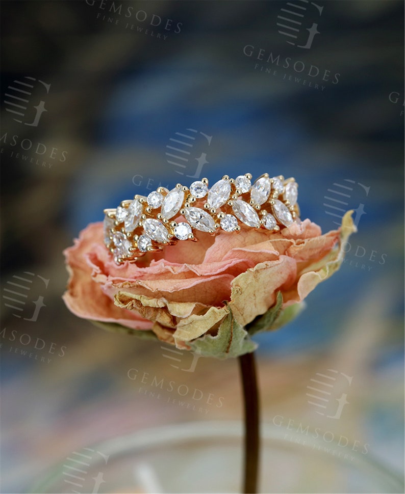 Puede incluir: Un anillo de oro con piedras preciosas claras de talla marquesa y redonda. El anillo est&aacute; sobre una rosa seca de color naranja claro. El fondo es una mezcla borrosa de azul, blanco y marr&oacute;n. El anillo es de Gemsodes Fine Jewelry.