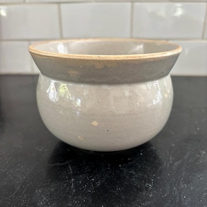 May include: A round, gray ceramic bowl with a slightly flared rim. The bowl has a smooth, matte finish and a light beige rim. The bowl is sitting on a black surface.