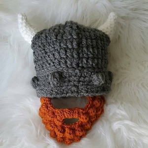 May include: A gray crocheted Viking helmet with white horns and an orange crocheted beard.
