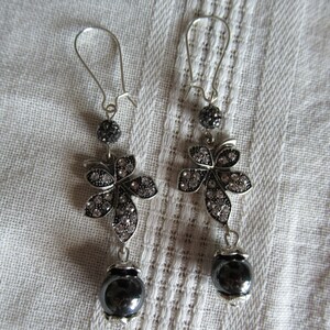 May include: A pair of silver dangle earrings featuring a black flower charm and a black bead. The flower charm is embellished with clear rhinestones.