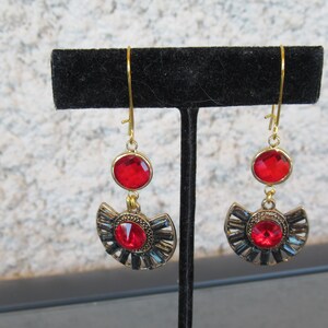 Può includere: Un paio di orecchini dorati con accenti di cristallo rosso. Gli orecchini presentano un design a forma di mezzaluna con un cristallo rosso più piccolo nella parte superiore.
