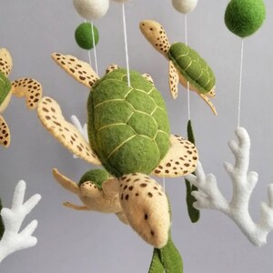 Puede incluir: Un m&oacute;vil de fieltro para beb&eacute; con tortugas marinas verdes y marrones, coral blanco y algas verdes que cuelgan de un anillo de madera. El m&oacute;vil tambi&eacute;n tiene bolas de fieltro blancas.