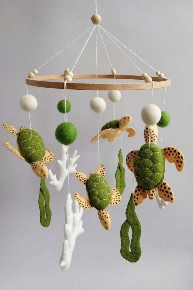 Puede incluir: Un m&oacute;vil de beb&eacute; de madera con decoraciones de fieltro de tortuga marina y coral. El m&oacute;vil incluye cuatro tortugas verdes y amarillas, dos piezas de coral blanco y bolas de fieltro verdes.